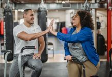 Two smiling people sitting on exercise bikes leaning over to give each other a high five.
