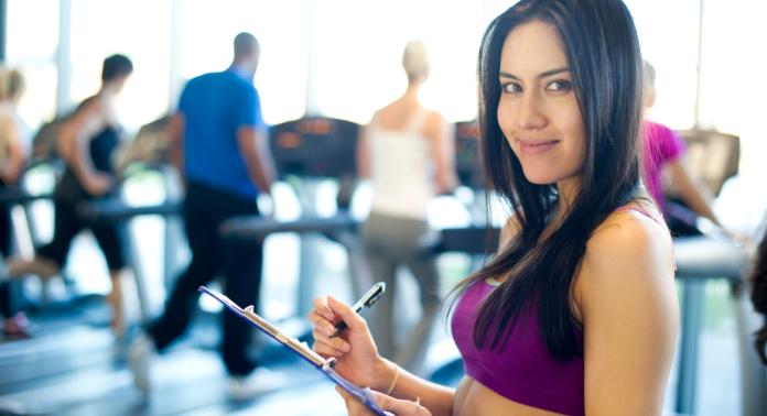 A woman smiling while holding a clipboard in a gym. In the background, two people are running out treadmills.