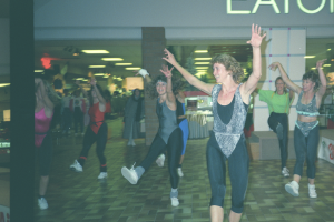 A grainy photo showing Christina Truscott leading a group fitness class in the 1980s. Everyone is wearing leotards and tights. Christina is holding her arms up in the air with a big smile on her face.