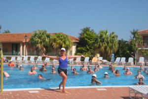 Christina Truscott smiling and posing with her arms outstretched. Behind her is a pool with about 20 participants in an aquatic fitness class. Palm trees, beach chairs and a clear blue sky are in the background.